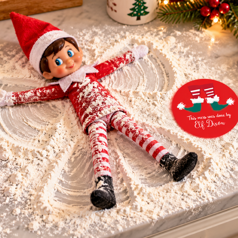 A Christmas-themed elf dressed as an elf lies on its back on a marble countertop covered in white flour, with a red circular sticker that reads 'This was you done by Elf Boss' nearby. The scene includes holiday decor such as a metal flour canister, a red cookie cutter, pine branches, and warm string lights.