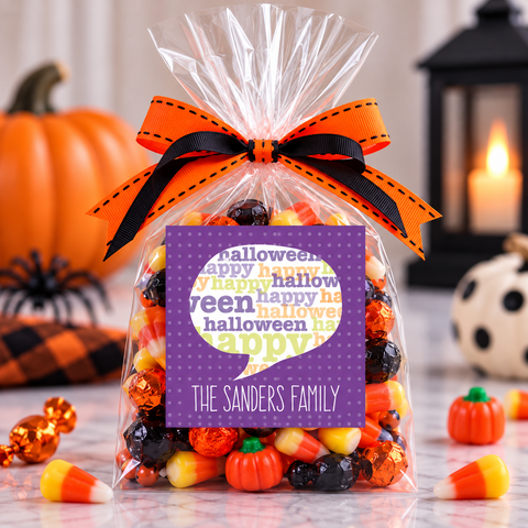 A clear cellophane gift bag filled with assorted Halloween candies, tied with an orange and black ribbon, and featuring a purple custom label that reads 'The Sanders Family' with 'halloween' in the background. The bag is styled on a marble surface with pumpkins, a candle, and candy corn in the background.