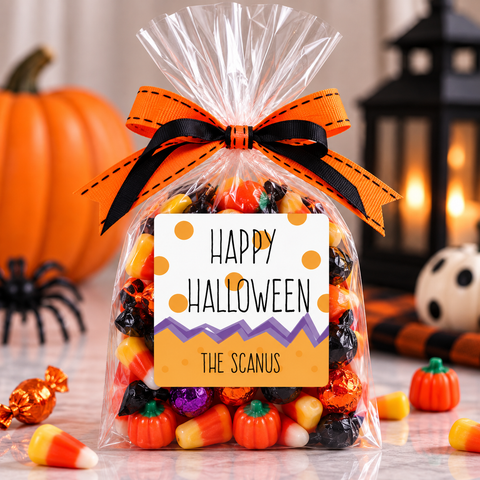 A clear cellophane gift bag filled with assorted Halloween candy, tied with an orange and black ribbon, and labeled 'Happy Halloween The Scanus.' Pumpkins, candy corn, a black spider decoration, and a lantern are visible in the background.