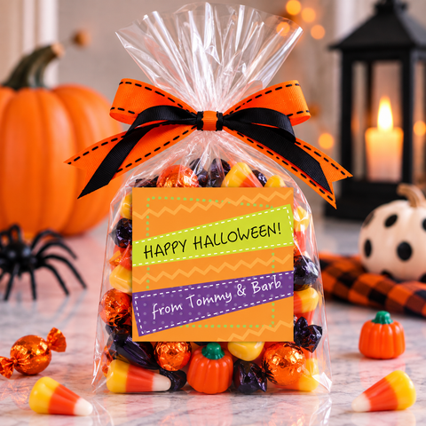 A clear cellophane gift bag filled with assorted Halloween candies, tied with an orange and black ribbon, and labeled 'Happy Halloween! From Tommy & Barb'. The bag is set on a marble surface with pumpkins, candy corn, a spider decoration, and a lit candle in the background.