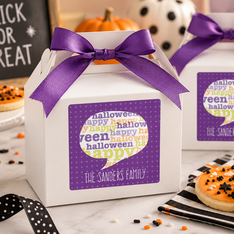 Two white Halloween-themed favor boxes tied with purple ribbons, decorated with a purple and white 'halloween happy family' label, set on a marble table with cookies, pumpkins, and a 'Trick or Treat' sign.