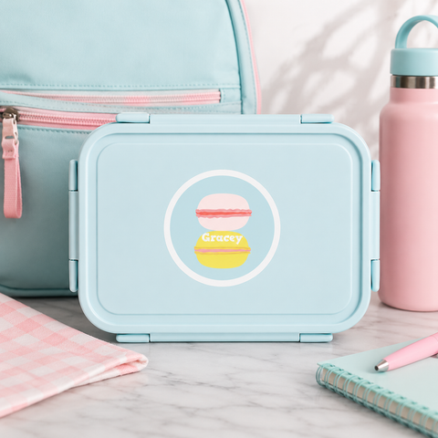 Light blue lunch box with a macaroi personalized label design, pink water bottle, and pink bag on a marble surface.