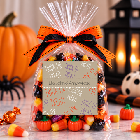 A clear cellophane gift bag filled with assorted Halloween candy, tied with an orange and black ribbon, and labeled 'Trick or Treat' for Ella, John, & Amy Wilcox. The bag is styled on a marble surface with pumpkins, a spider decoration, and a lantern in the background.