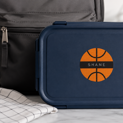 Navy blue lunch box with basketball design and persoalized with your name, placed on a marble surface with a backpack and water bottle in the background.