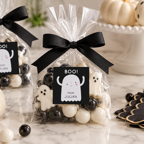 Three clear cellophane favor bags tied with black ribbons and printed with ghost labels reading 'BOO! FROM JULIAN,' filled with black-and-white candies, arranged on a marble surface with pumpkins, straws, and black-and-gold 'Trick or Treat' cards.