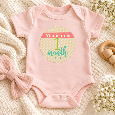 Onesie flat lay with a personalized first month baby girl milestone sticker