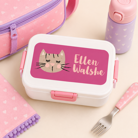 Pink lunch box with cat illustration with  pink and purple accessories on a beige background.