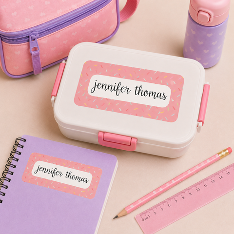 Personalized pink confetti waterproof name label on a white bento lunch box and lavender notebook with matching pastel school supplies.
