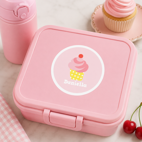 Personalized cupcake waterproof name label on a pastel pink lunch box with matching pink water bottle, cupcake-themed accessories, and coordinated pastel props.