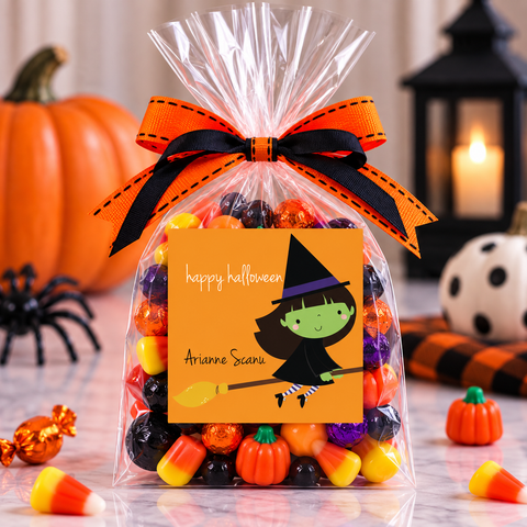 A clear cellophane gift bag filled with assorted Halloween candies, tied with an orange and black ribbon, and featuring a personalized printed 'Happy Halloween' label The bag is styled on a marble surface with pumpkins, a spider, candy corn, and a lantern in the background.